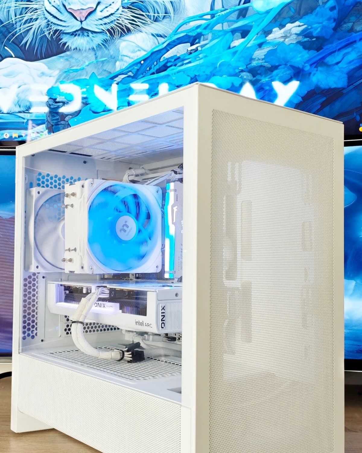 Bílý herní počítač NZXT H3 Flow s modře podsvíceným chladičem a grafikou Intel Arc B580 před monitorem s bílým tygrem